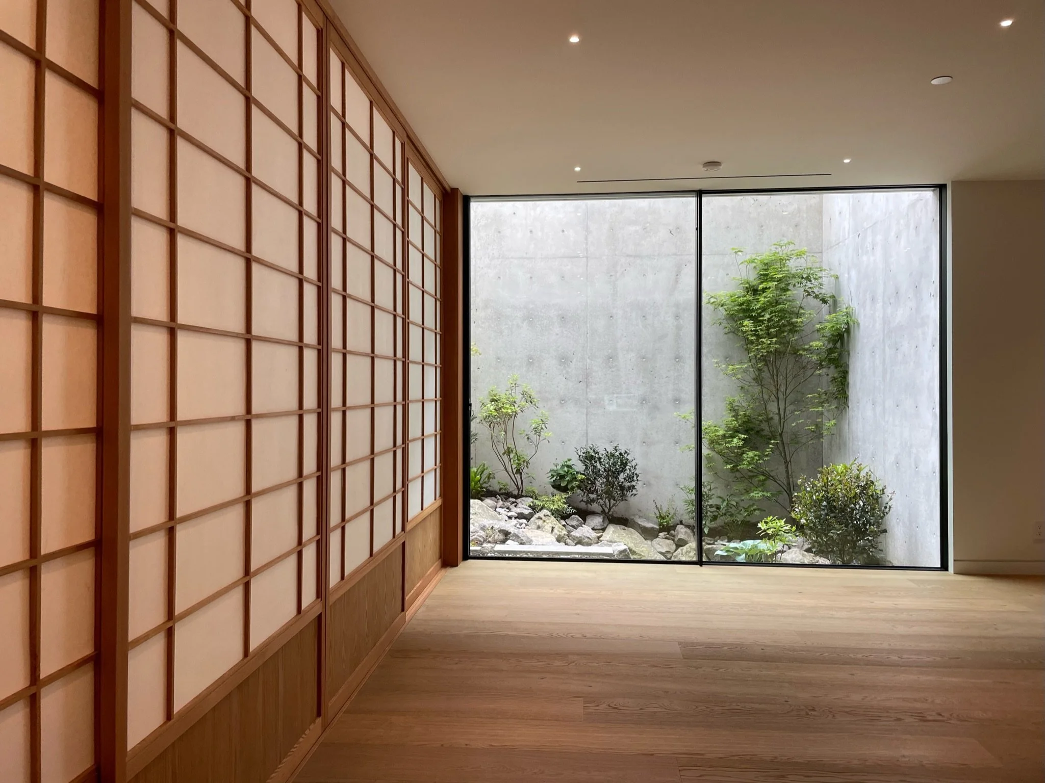 Minimalist interior with wooden flooring, shoji-style sliding wooden doors on the left, and a large floor-to-ceiling window revealing a small outdoor garden with rocks, green plants, and trees.