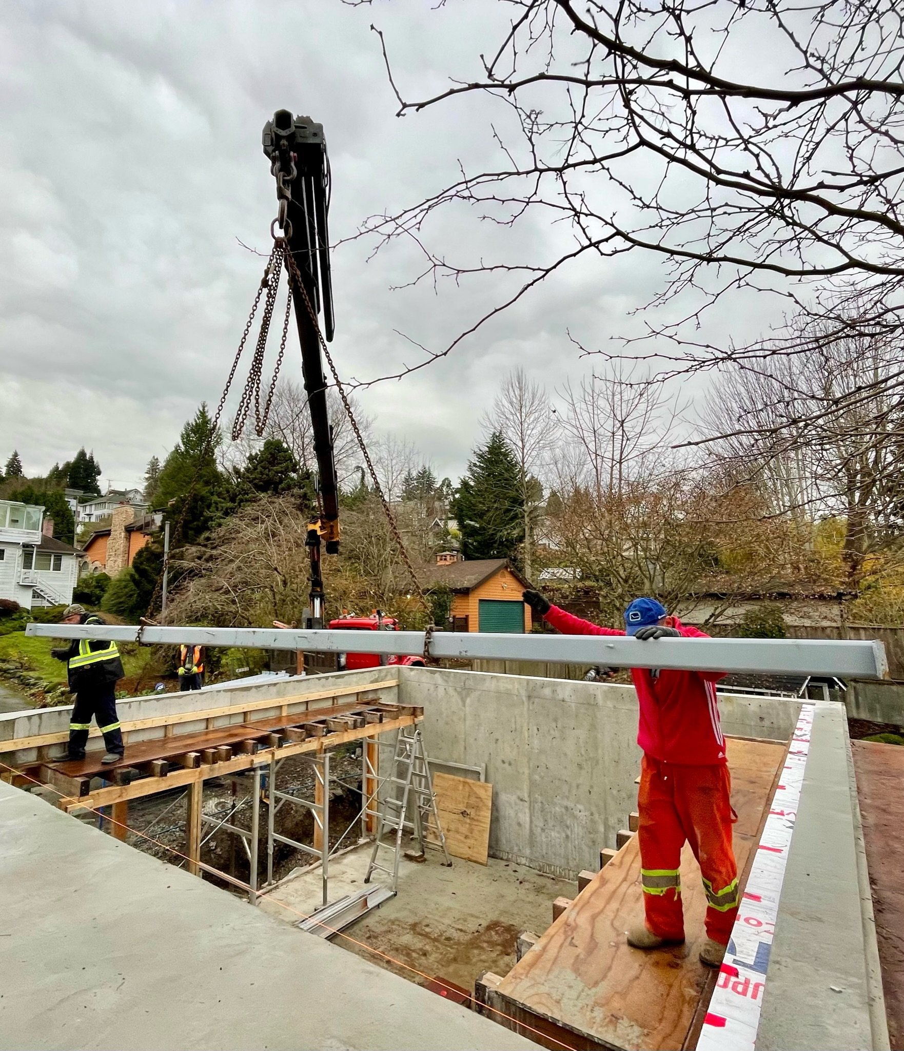 Construction workers are lifting and positioning a large steel beam at a building site during overcast weather, with trees and houses in the background.