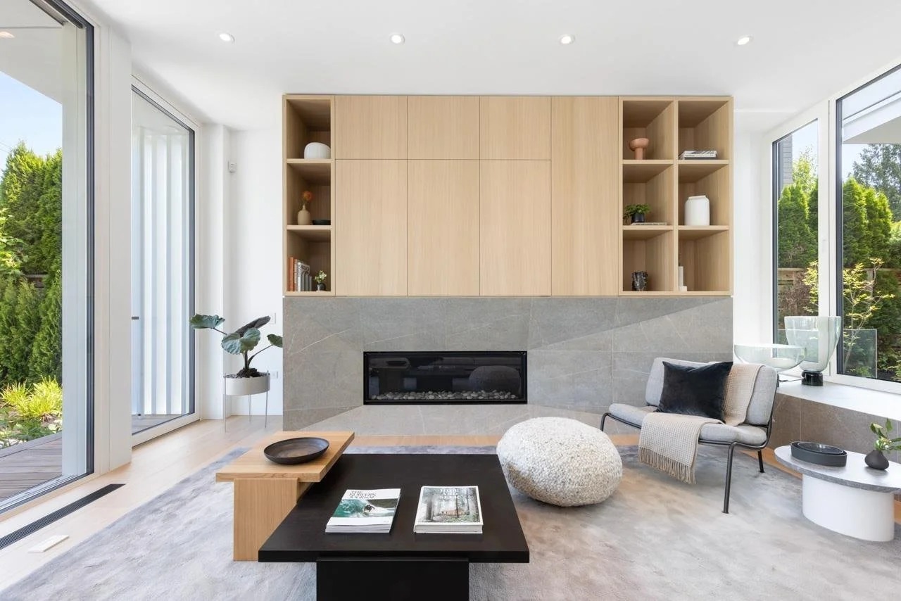 Modern living room with a built-in fireplace, light wood shelving, large windows showing a garden, a gray armchair with a black pillow, a round white ottoman, a black coffee table with magazines, and decorative items.