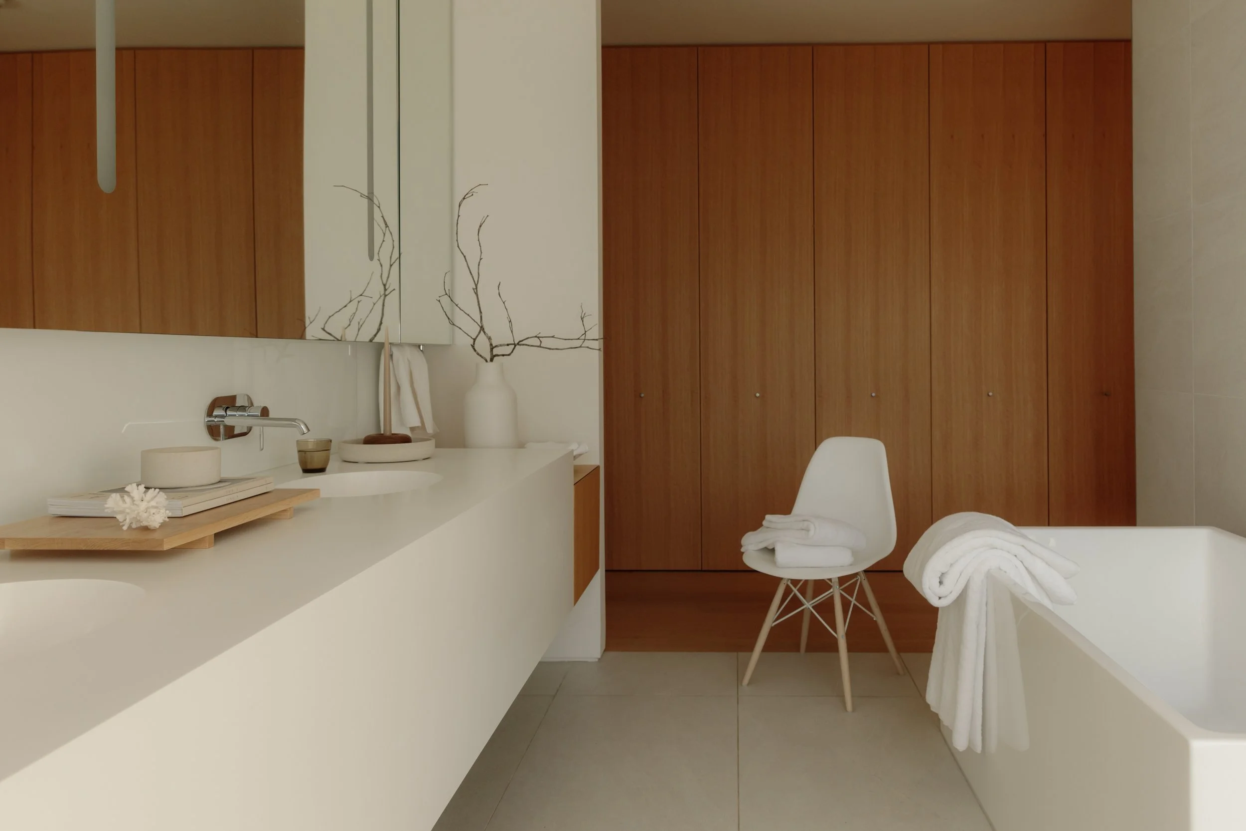 Minimalist bathroom with white vanity, wooden cabinets, a white chair with towels, bathrobe on edge of tub, and decorative vase with branches.