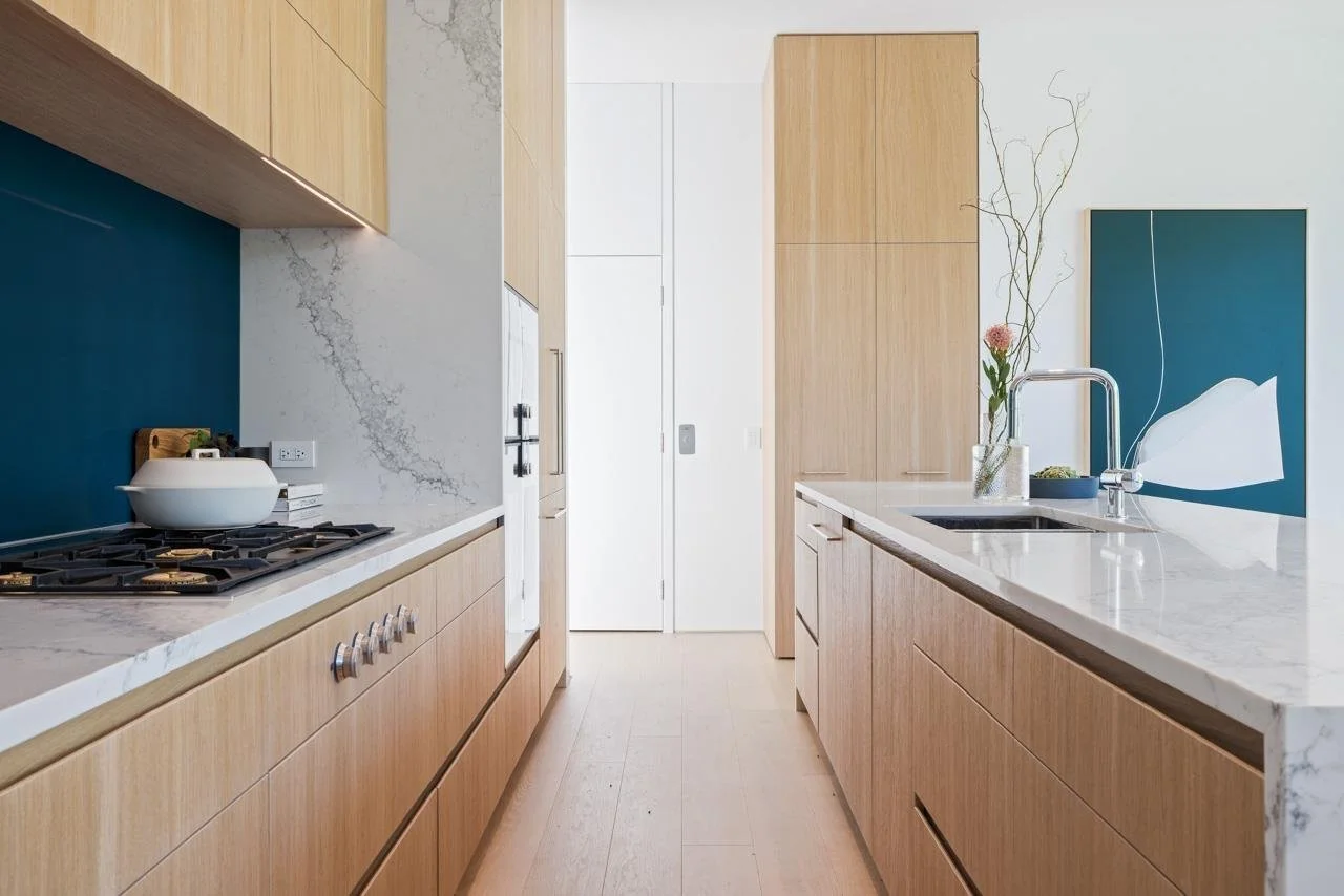 Modern kitchen with light wood cabinets, white marble countertops, a blue backsplash, and a vase with flowers on the sink