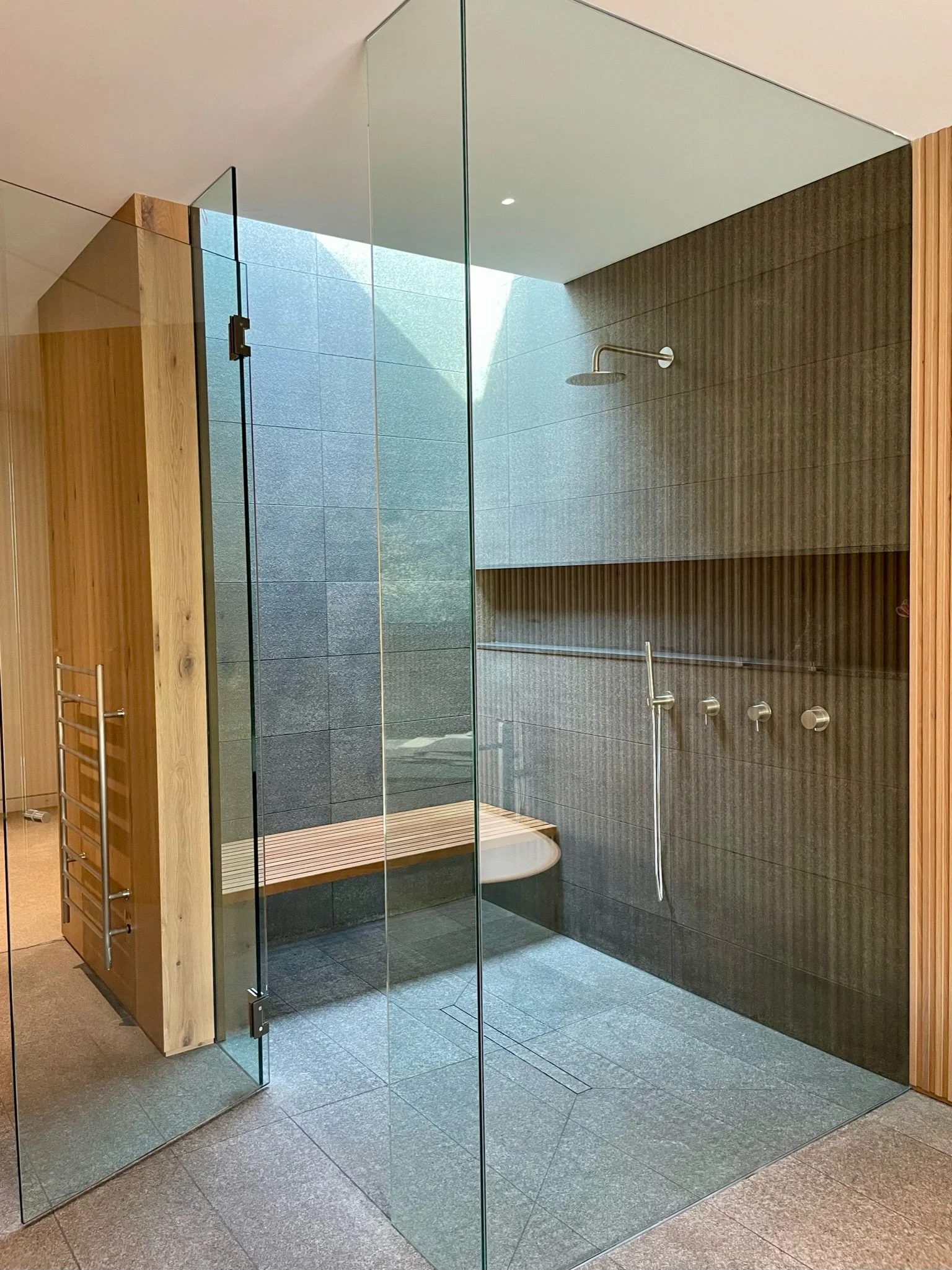Modern indoor shower with glass doors, dark gray tiled walls, a ceiling-mounted showerhead, a wooden bench, and a horizontal grab bar.