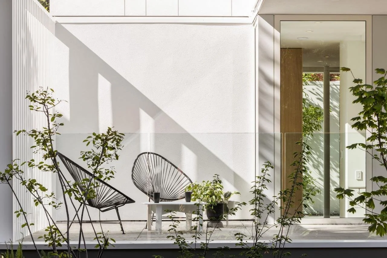 Modern outdoor patio with two black wire chairs, a small white table, and potted plants, in front of a white wall with shadows, adjacent to a glass door leading into a room.