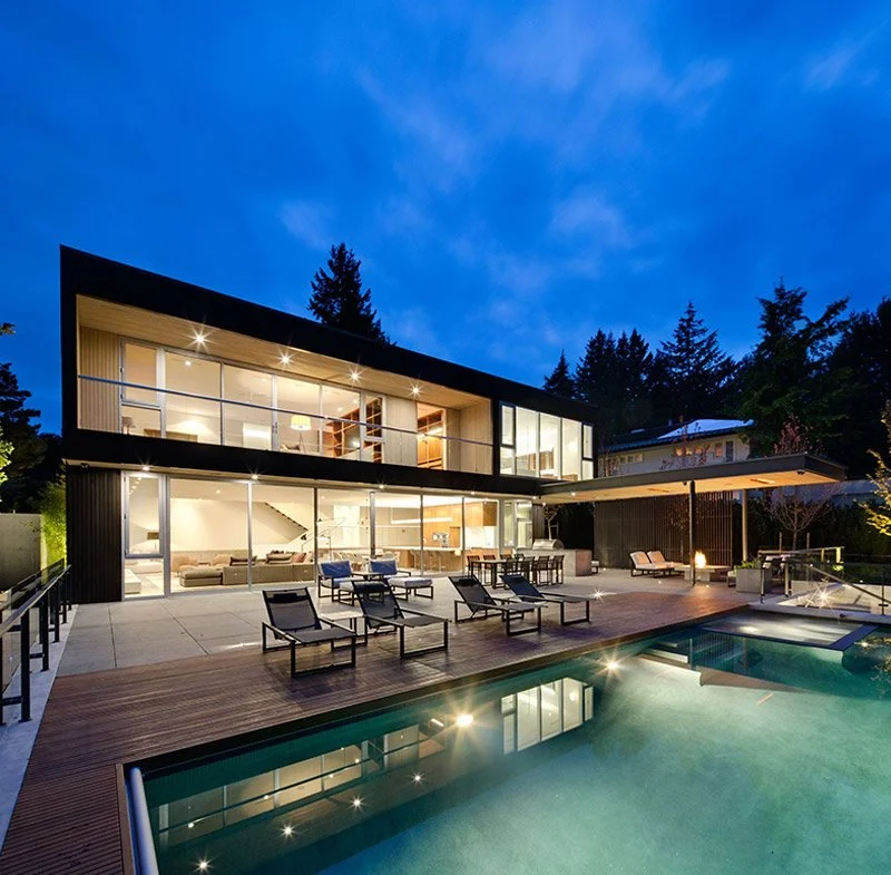 Modern two-story house with large glass windows, outdoor lounge chairs, a pool, and surrounding trees, during evening.