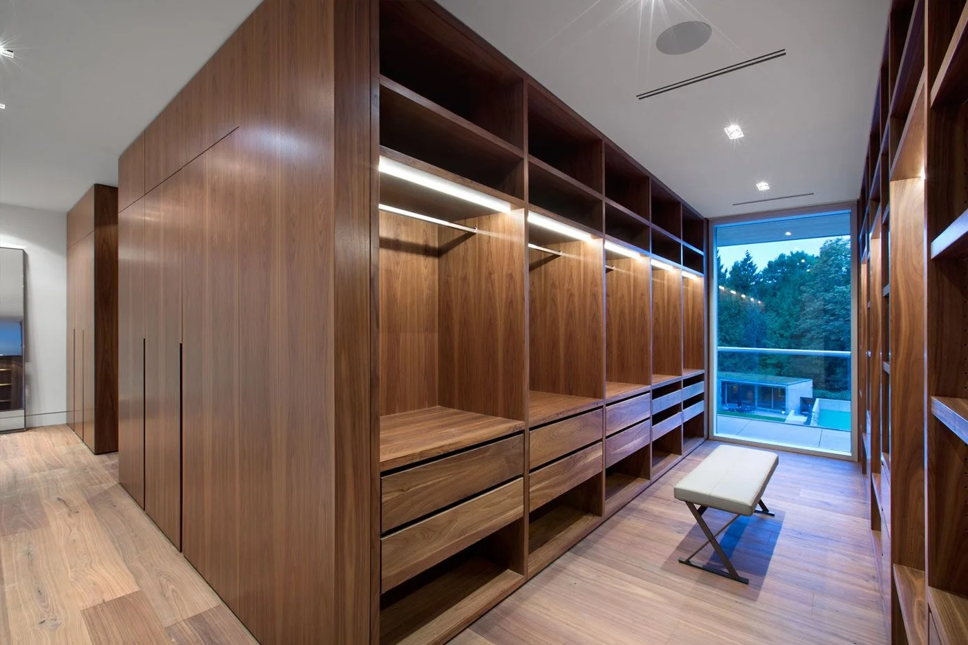 Empty wooden walk-in closet with built-in shelving, drawers, a cushioned bench, and a large window showing outdoor trees.