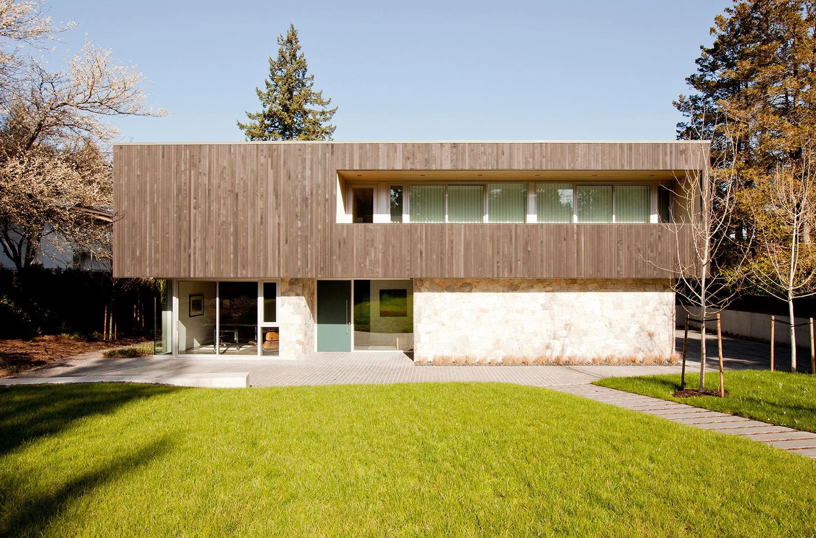Modern two-story house with wooden siding on the upper level and stone on the lower level, surrounded by a green lawn and leafless trees.