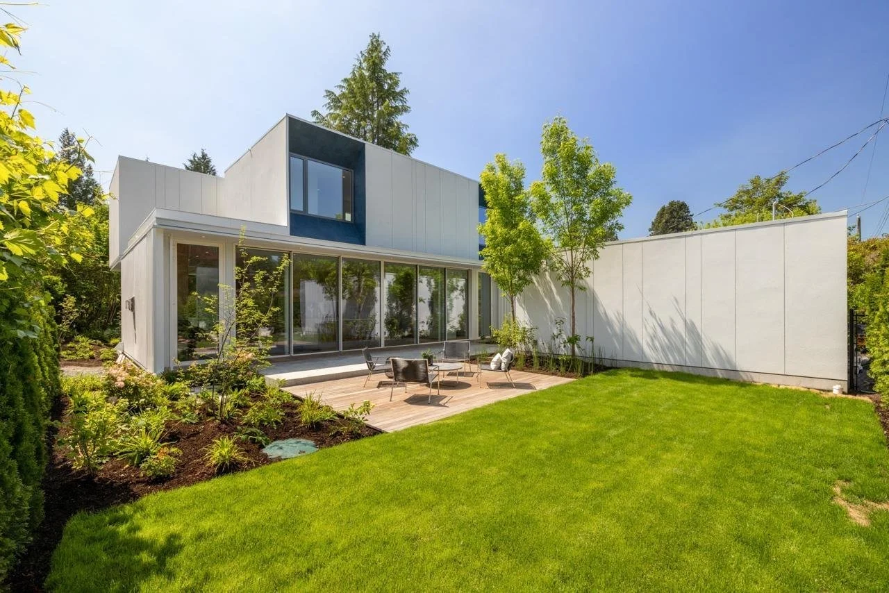 Modern white house with large glass sliding doors, outdoor patio with chairs and table, green lawn, and trees under a clear blue sky.