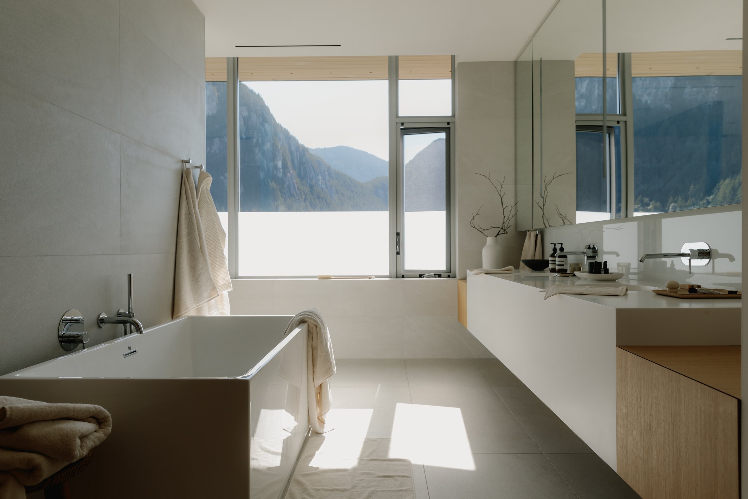 Modern bathroom with large windows showcasing mountain scenery, bathtub with towels, and minimalist vanity with mirror and decor.