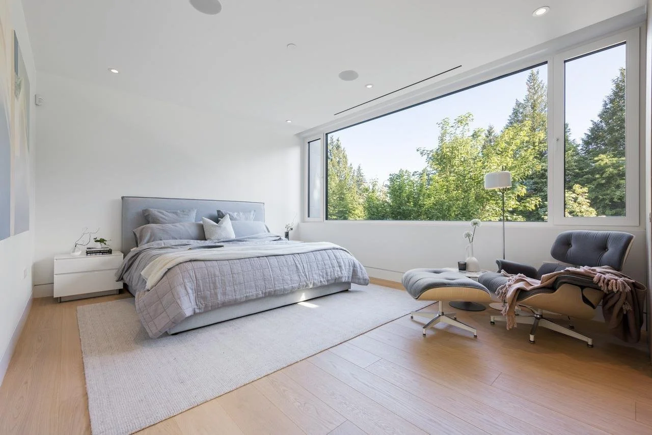 Minimalist bedroom with large window view of trees, gray bed with pillows, white nightstand, and gray lounge chair with matching ottoman.