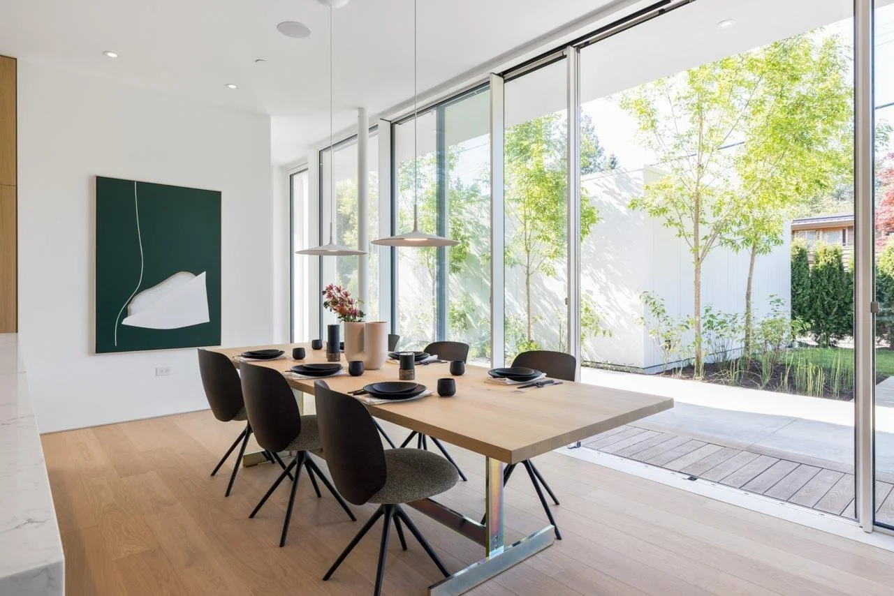Modern dining room with a wooden table set with black plates and cups, surrounded by six black chairs, large glass sliding doors opening to a garden with trees, bright natural light, and contemporary artwork on the white wall.
