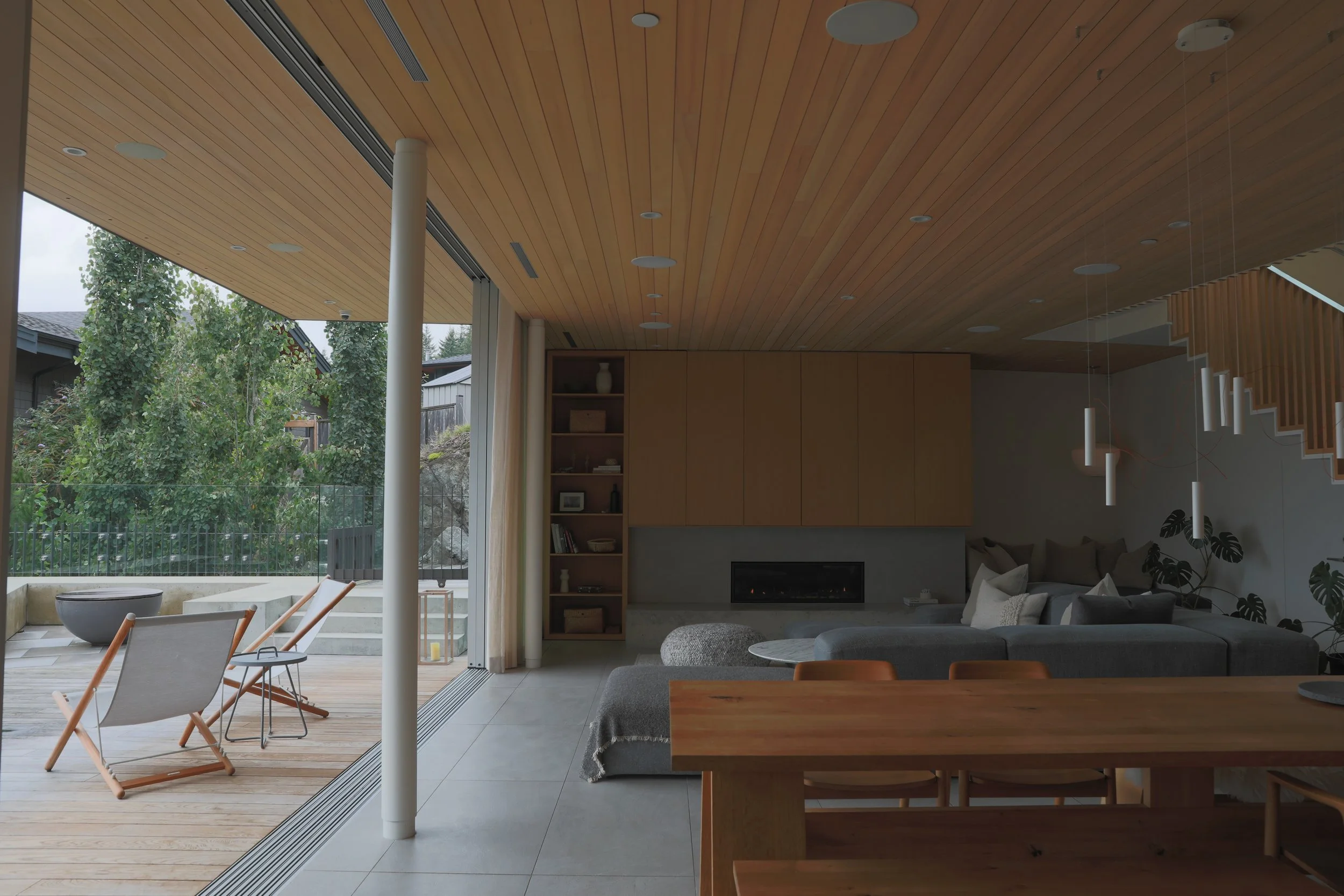 Modern living room with large glass sliding door open to outdoor deck with lounge chairs, visible trees, and neighboring houses. Interior features a wooden ceiling, built-in shelves, fireplace, gray sectional sofa with pillows, a gray ottoman, a wood