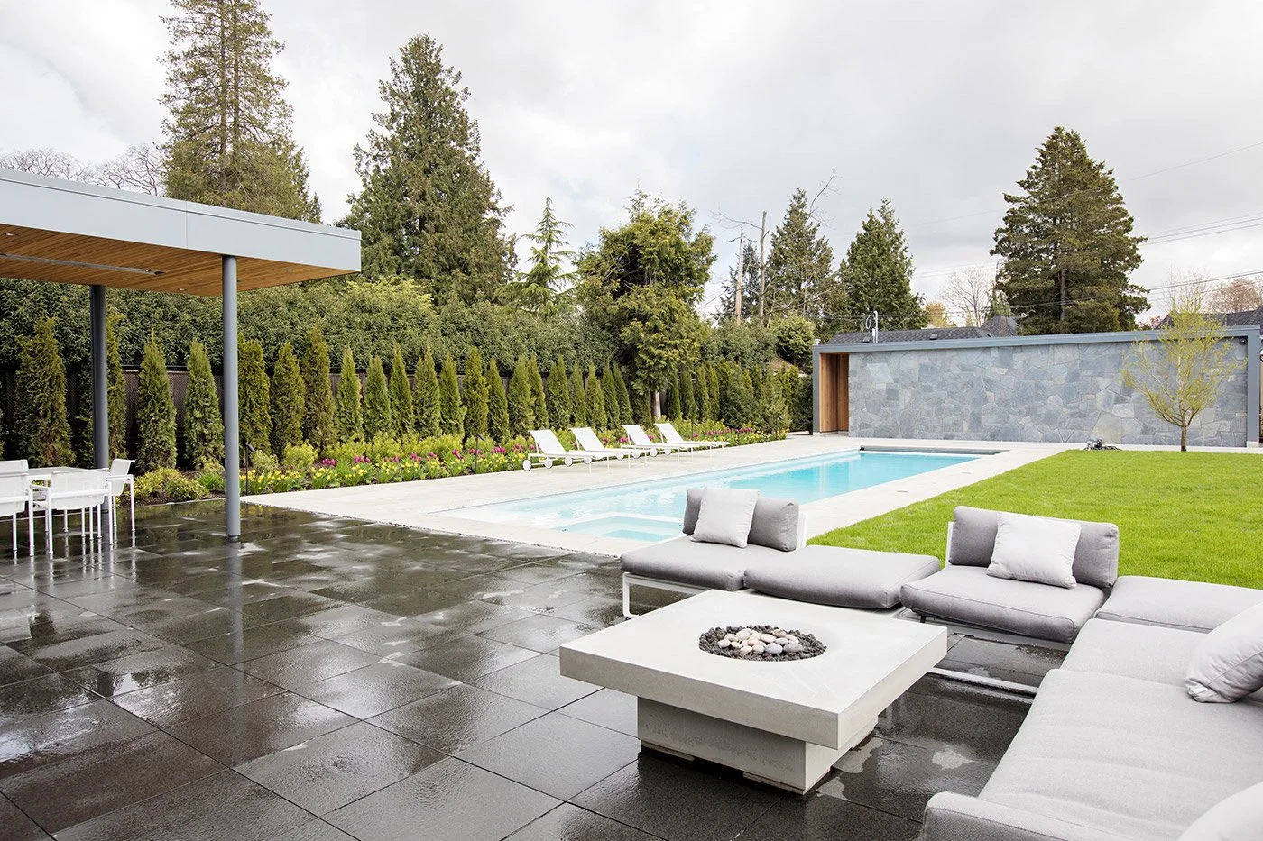 Modern backyard with swimming pool, gray patio furniture with cushions, green lawn, and tall trees in the background.