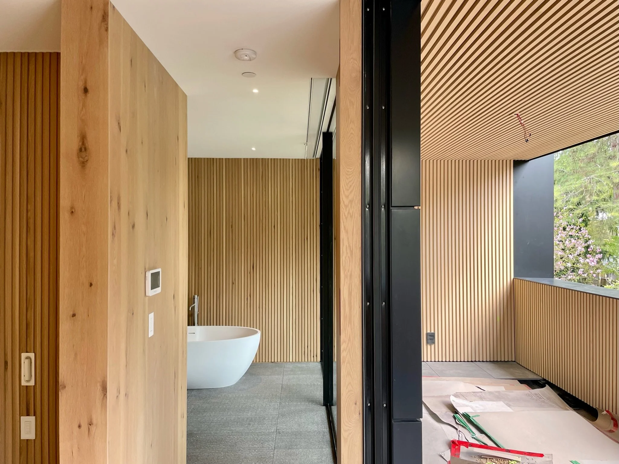 Interior of a modern room with wooden paneling on the walls and ceiling, a white freestanding bathtub and a balcony with trees outside.
