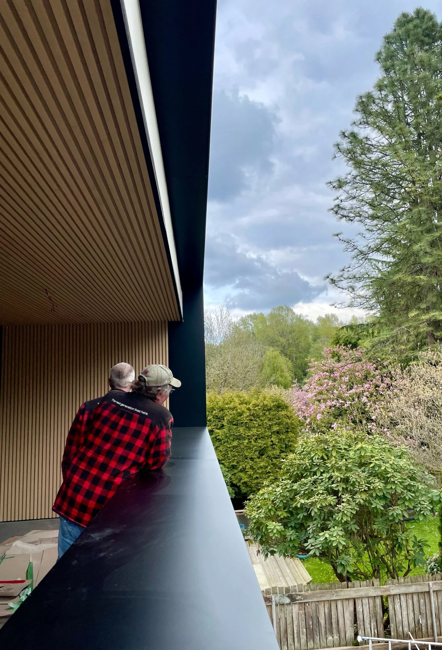 Two men leaning on a black railing on a balcony, looking out at trees and flowering bushes in a backyard, with a cloudy sky overhead.