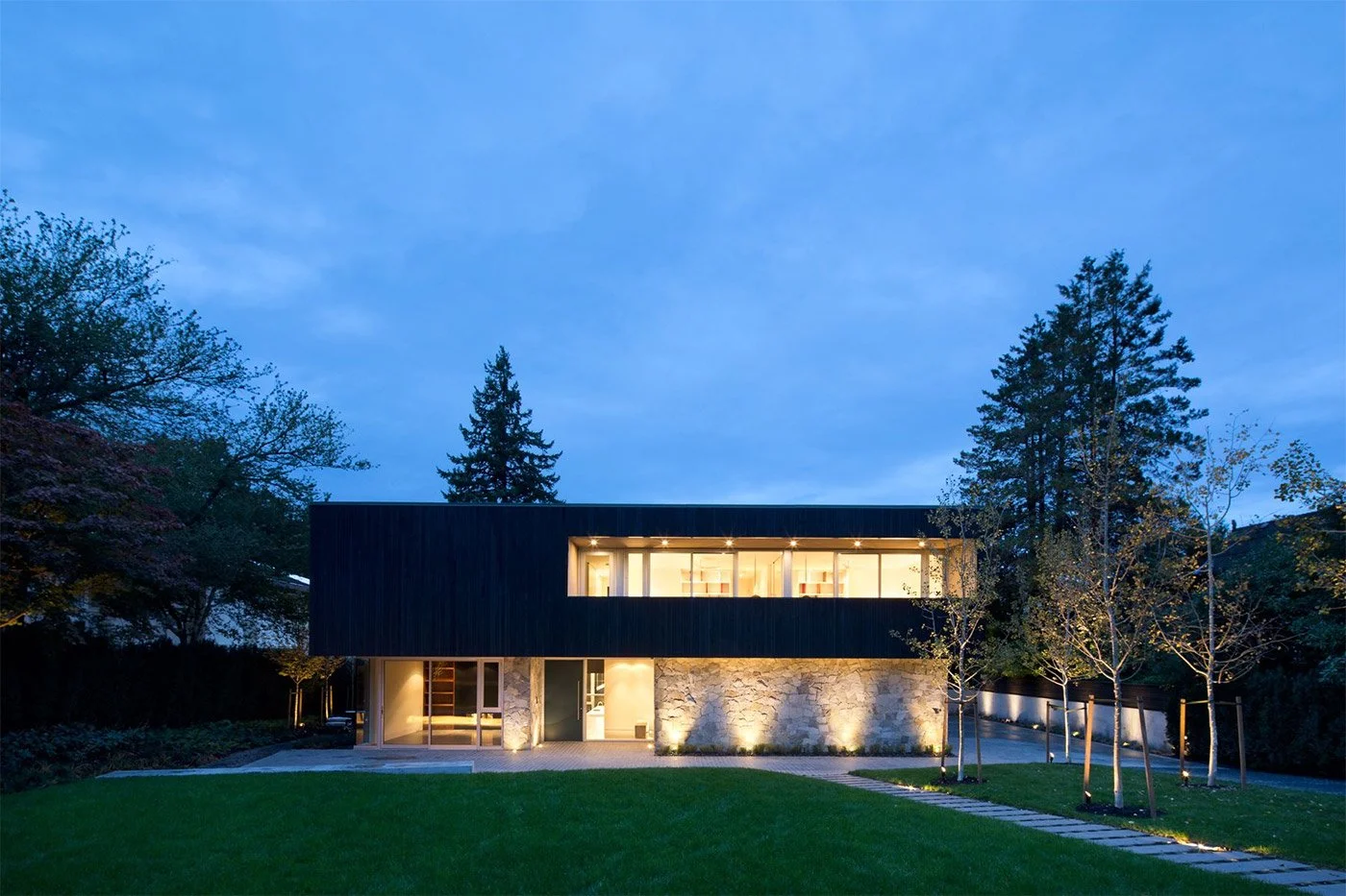 Modern two-story home with large windows and stone exterior, illuminated at dusk, surrounded by a lawn and small trees.