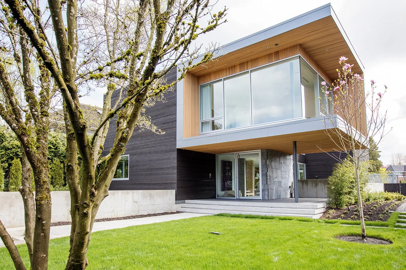 Modern two-story house with black, wood, and stone exterior, large windows, a front porch with steps, and a well-maintained lawn and trees in the yard.