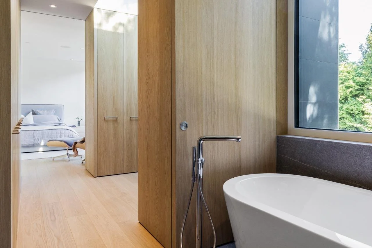 Modern bedroom and bathroom with wooden walls and floors, a bathtub by a large window overlooking green trees, and a bed with white bedding in the background.