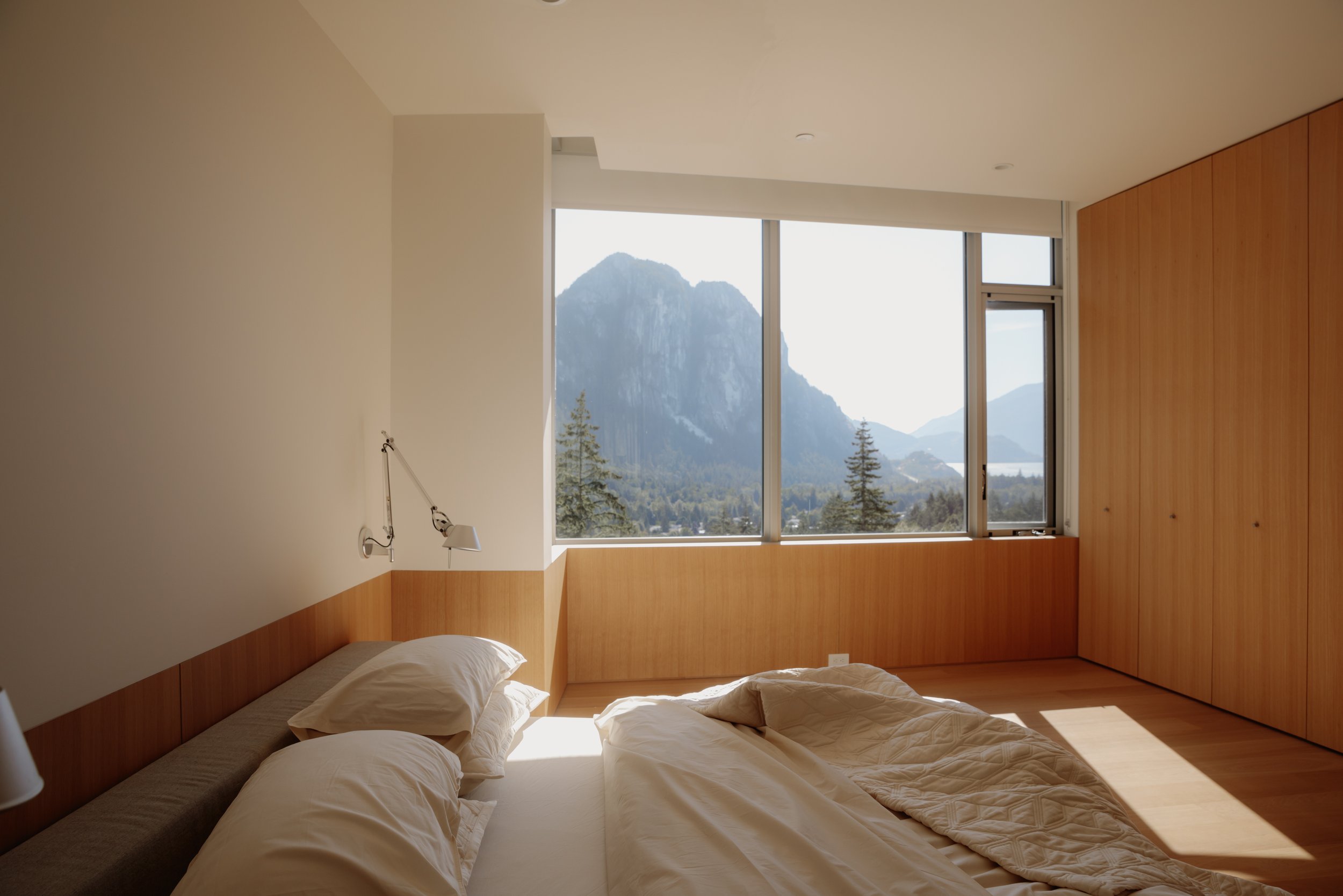 A bedroom with a bed, white pillows, and beige bedding, featuring large windows showing mountains and trees outside, with sunlight streaming in.