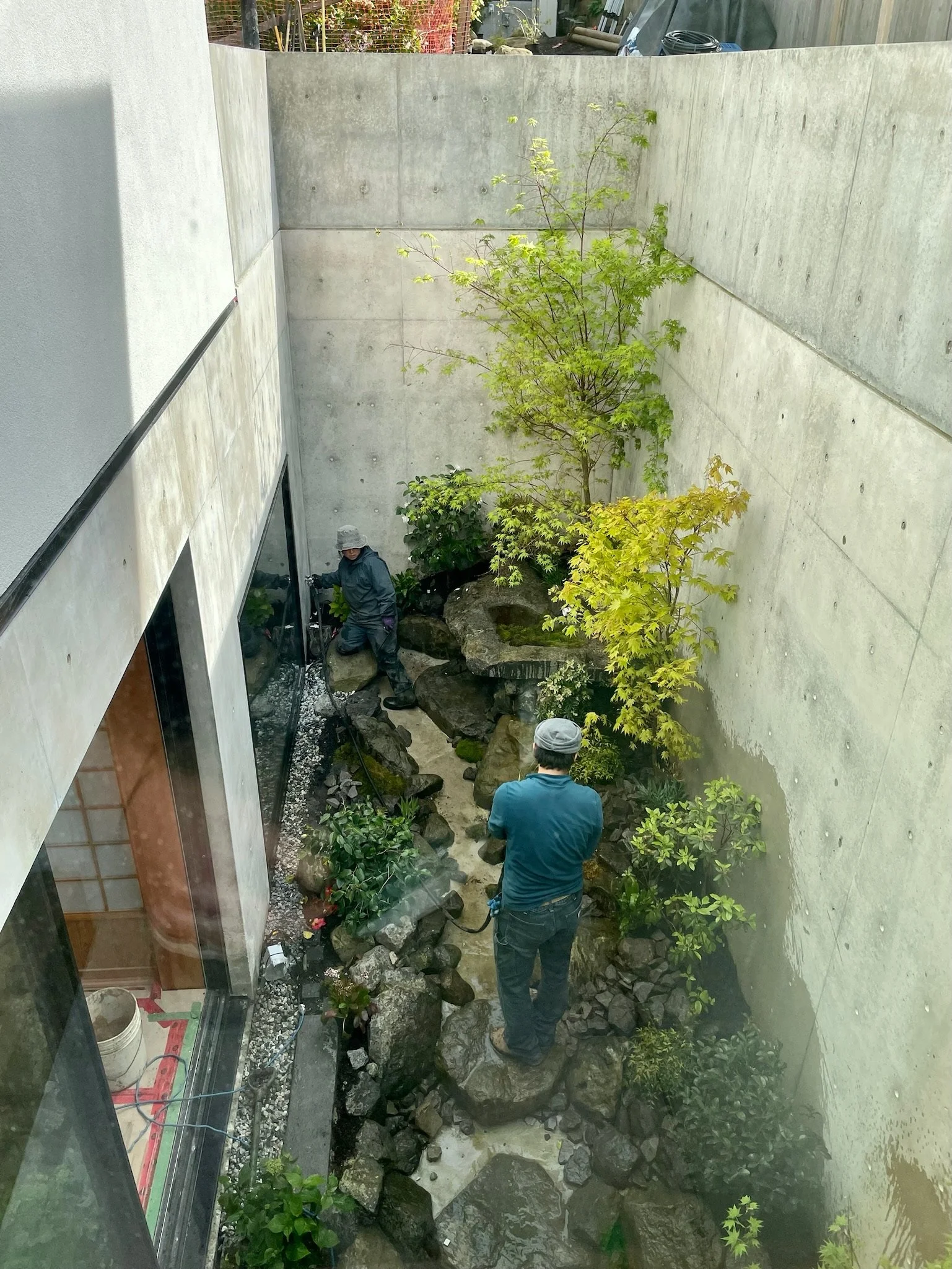 Two men are working on a small landscaped garden with rocks, plants, and a tree, enclosed by concrete walls, seen from an overhead perspective.