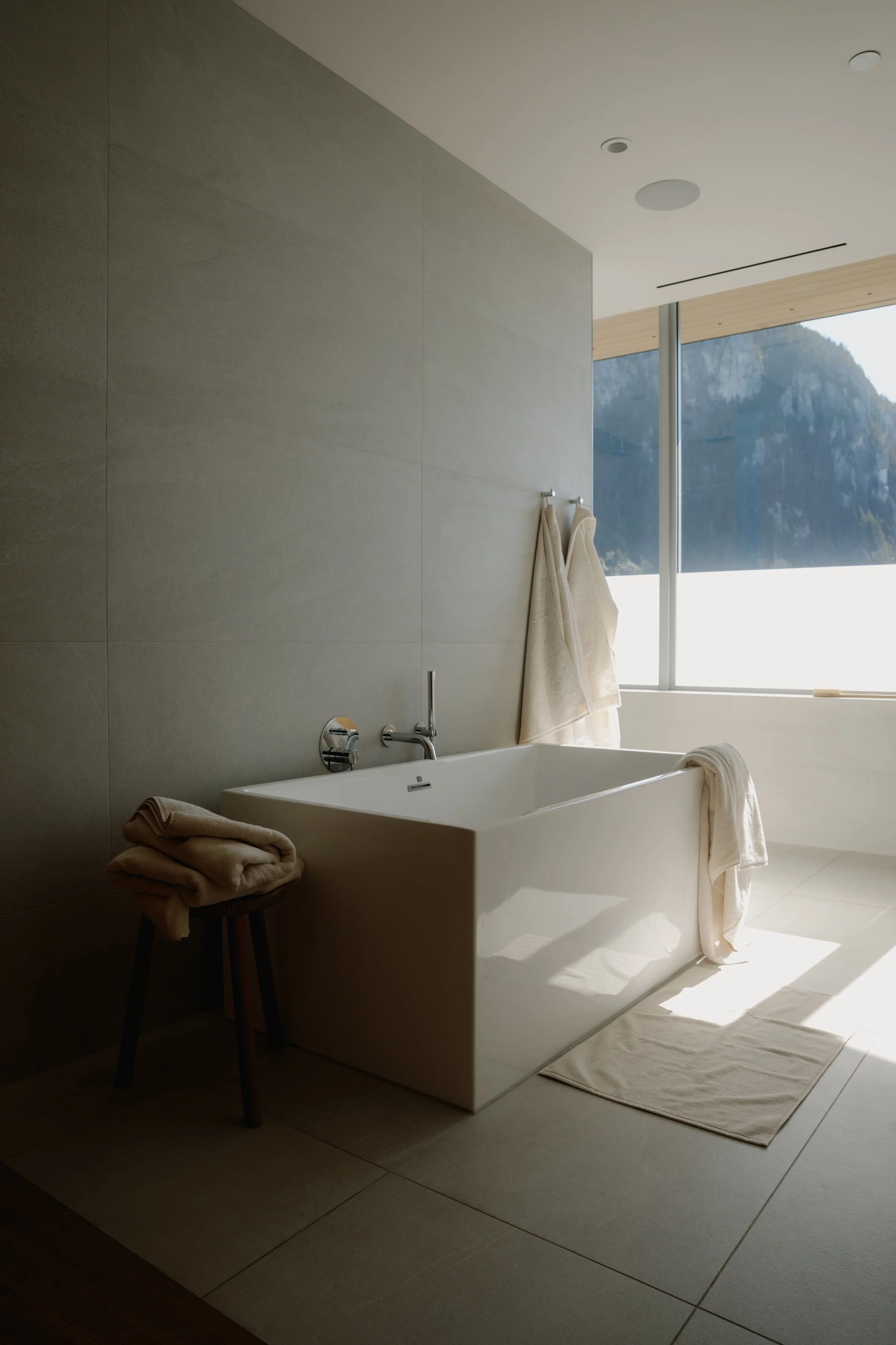 Modern bathroom with a white bathtub, beige towels on a stool, and a large window showing mountains outside.