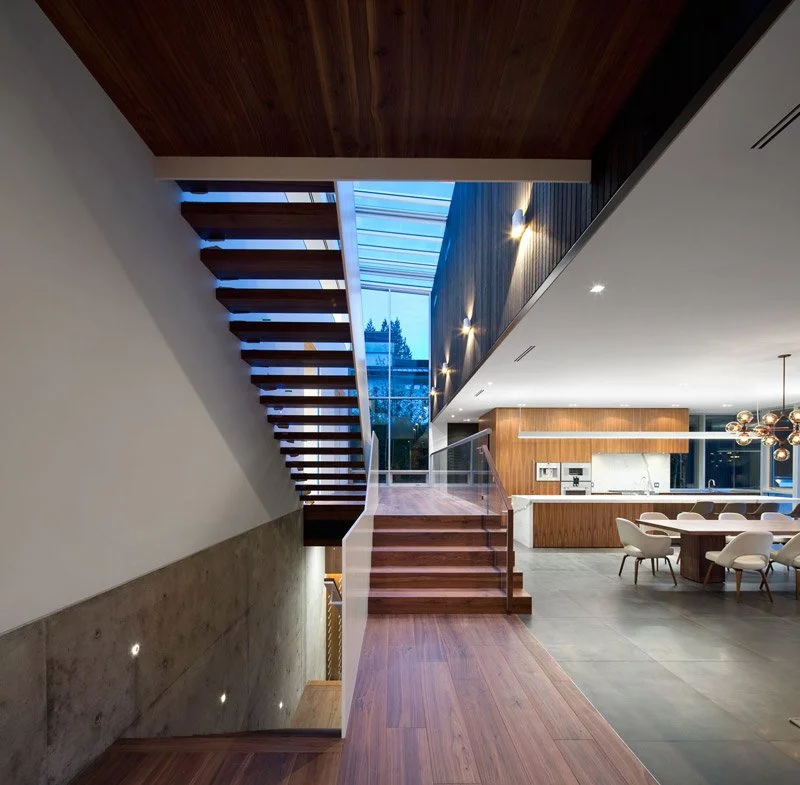 Modern interior of a home with a floating staircase, large glass window, open kitchen with wooden cabinets, dining area with a large table and white chairs, pendant lighting, and wooden ceiling accents.