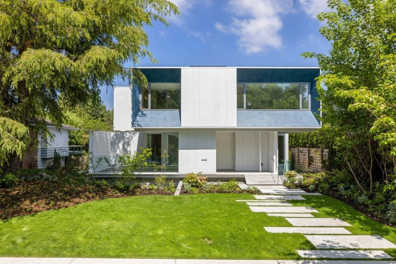 Modern two-story house with large windows, white exterior, and a well-maintained front yard with a stepping stone pathway, surrounded by lush green trees and lawn.