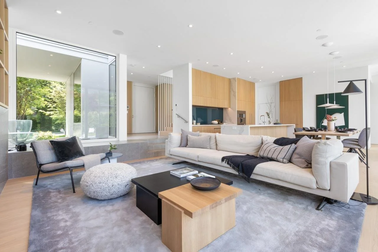 Modern open-concept living room with large white sofa, gray armchair, round textured pouf, black and wooden coffee tables, and a gray area rug. In the background, there is a kitchen with wooden cabinetry, a marble island, and a dining table with chai