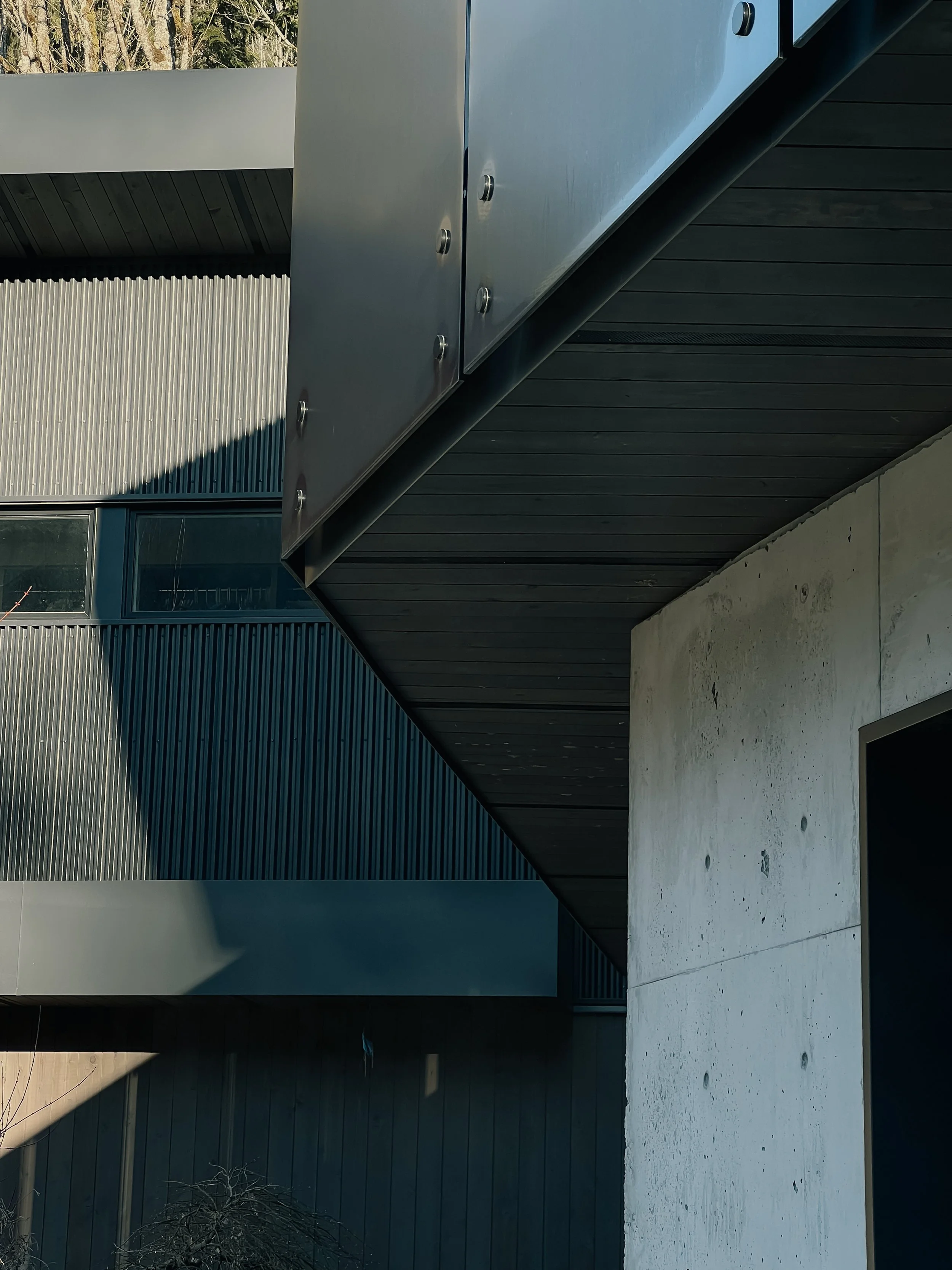 Close-up of modern building exterior with concrete wall, black metal roof soffit, and gray metal paneling with rivets, featuring a window reflecting the sky.