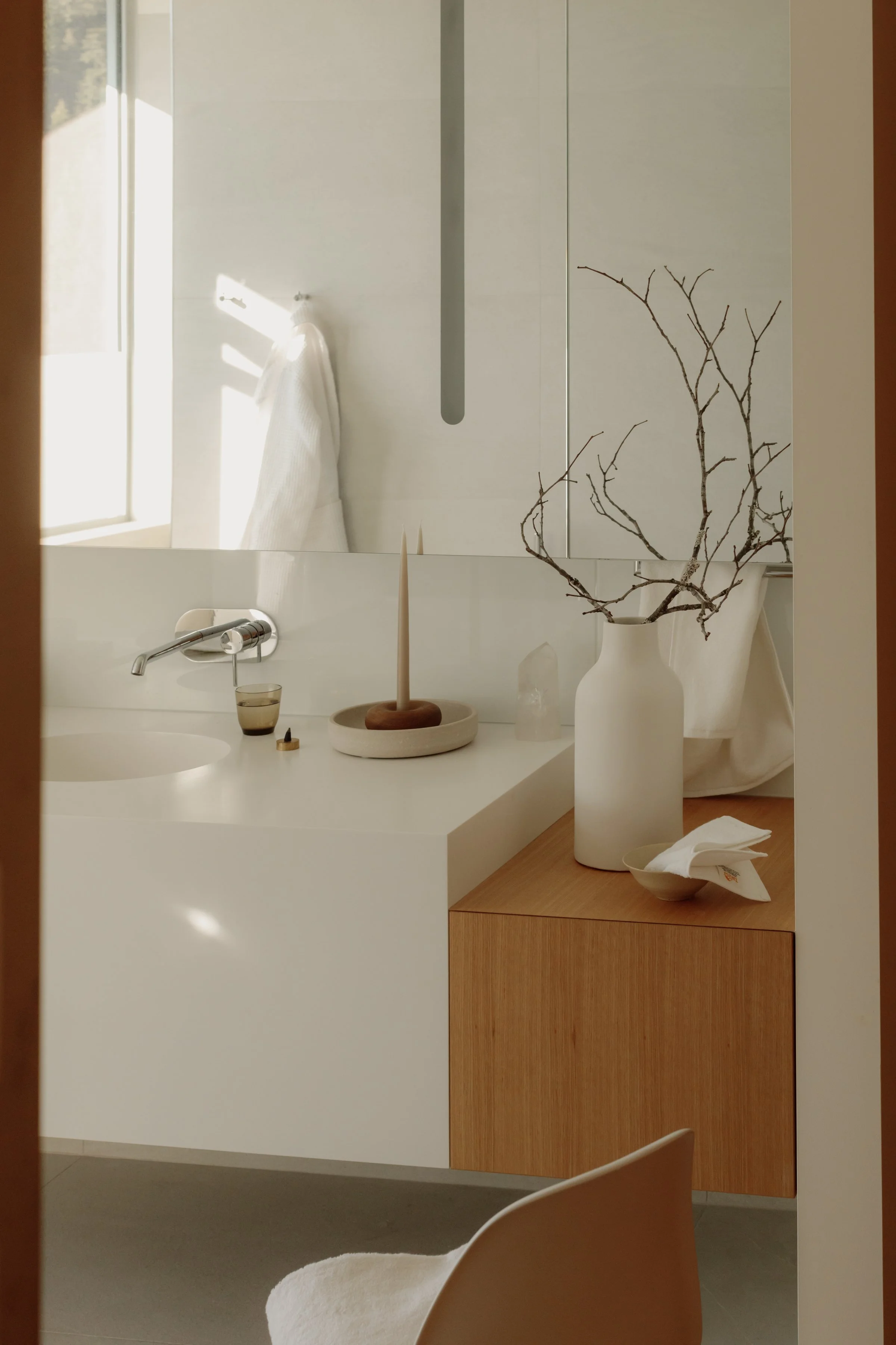 A modern bathroom with a white vanity, a mirror, a beige countertop, and decorative items including a large white vase with bare branches, a candle, a small bowl with napkins, and a glass of water.