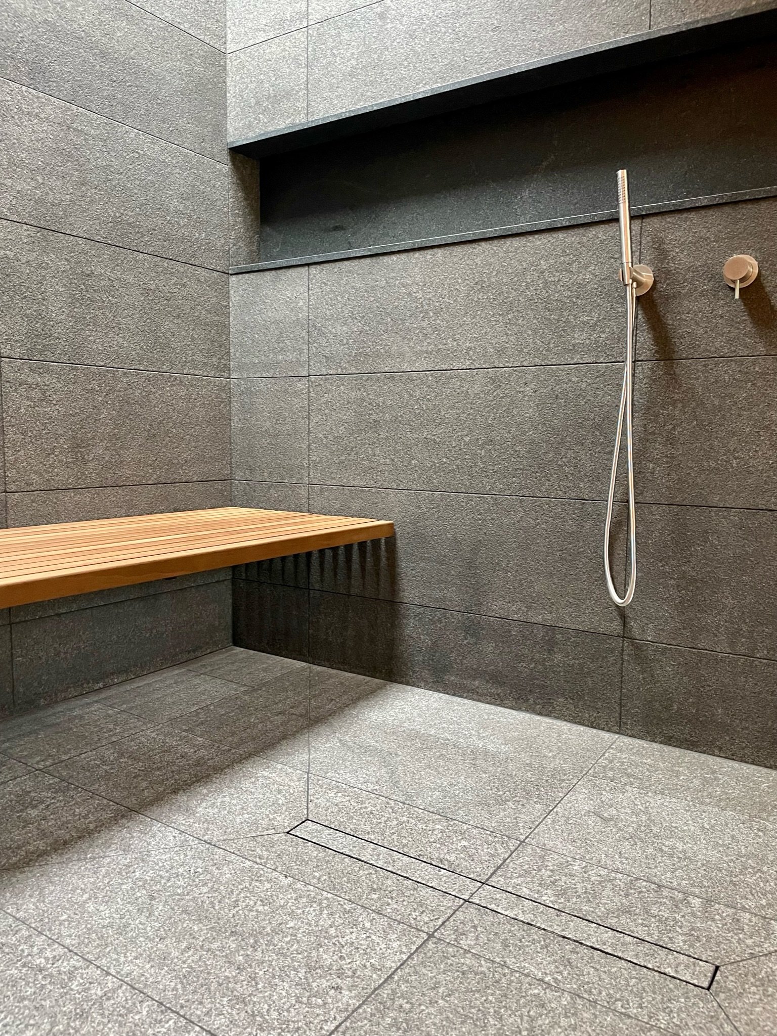 Modern shower area with gray tiled walls and floor, a wooden bench on the left, and a silver handheld showerhead mounted on the right wall.