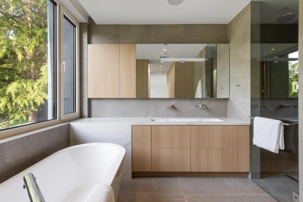 Modern bathroom with large window, freestanding bathtub, wooden vanity with marble countertop, large mirror, and walk-in shower with glass wall.