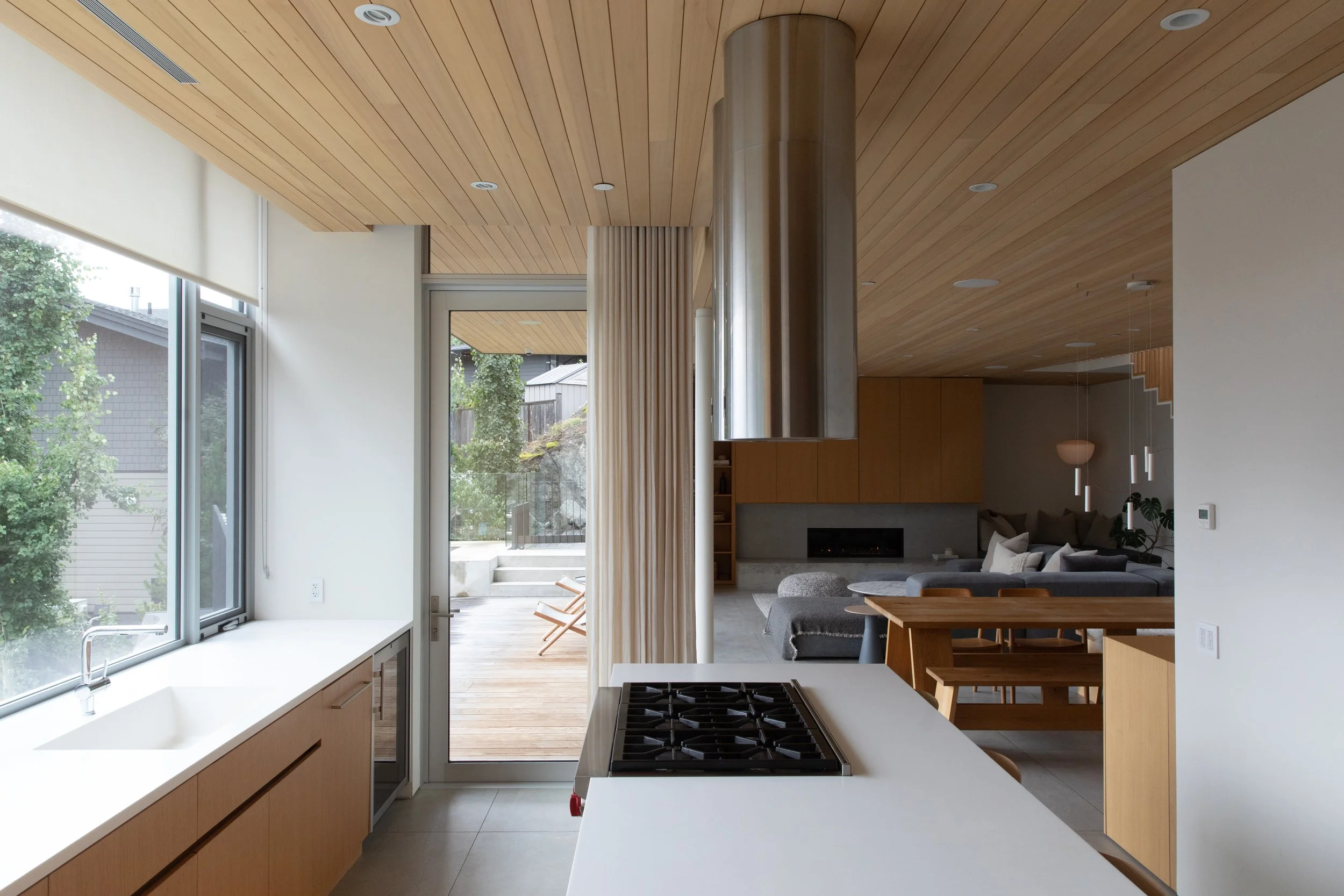 Modern open-concept kitchen and living room with wooden ceiling, large windows, and sliding glass door leading to an outdoor deck with chairs.
