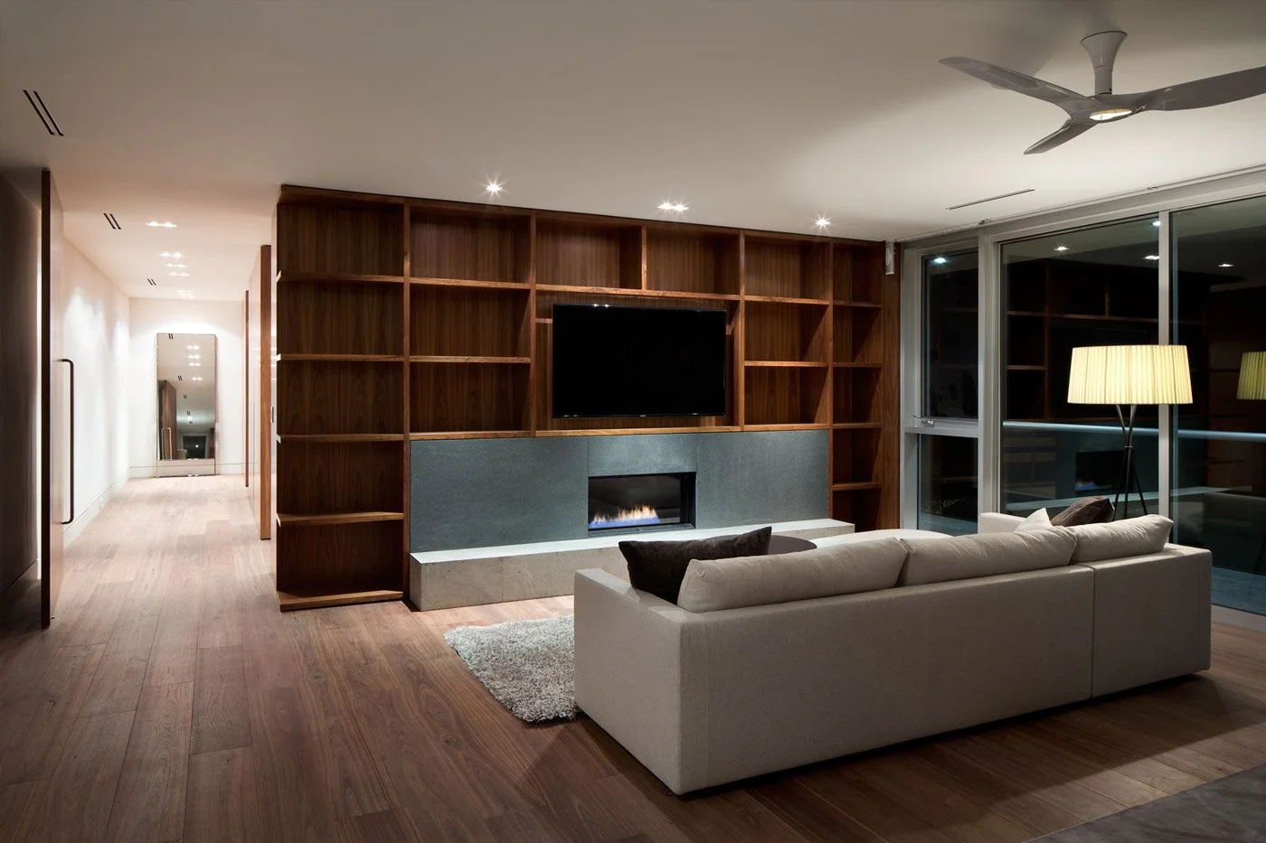 Modern living room with wooden built-in bookshelf, wall-mounted flat screen TV, gas fireplace, white sofa with black pillows, floor lamp with beige shade, large glass windows at night.