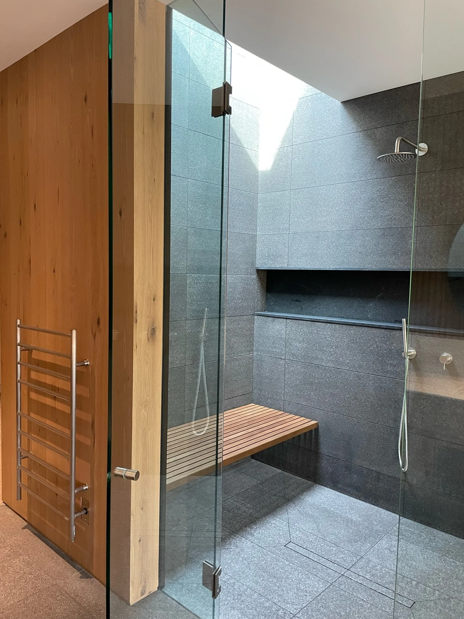 Modern shower area with glass doors, grey tiled walls and floor, a wall-mounted rain shower head, a built-in shelf, and a wooden bench. There is also a towel warmer on the adjacent wall.