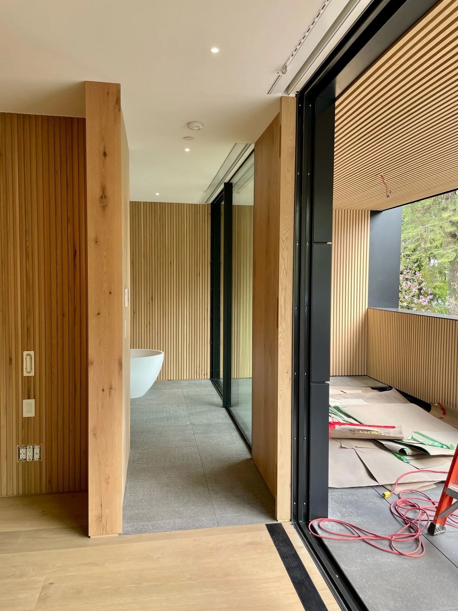 Interior view of a modern room with wooden paneling, a partially visible white bathtub, and large sliding glass doors leading to an outdoor balcony with construction materials and tools.