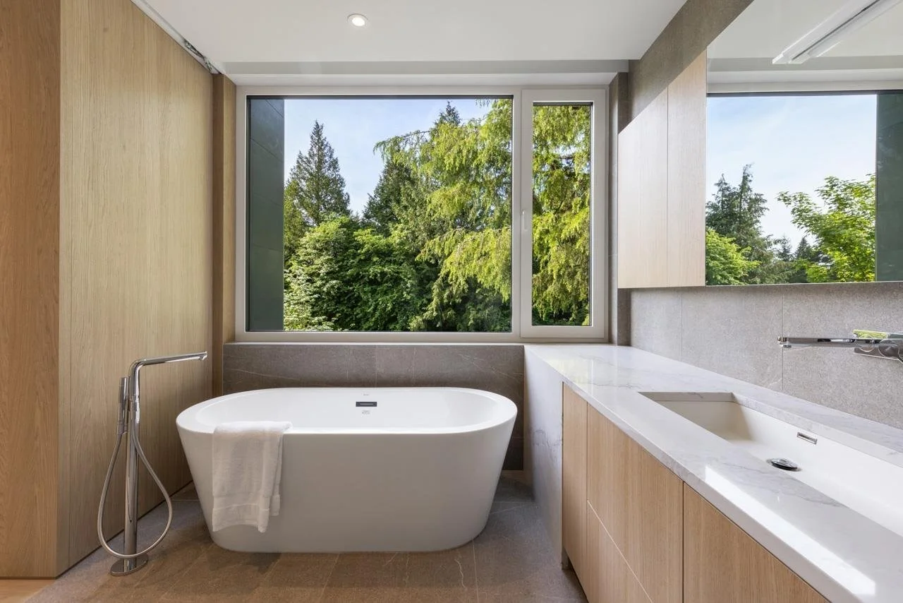 Modern bathroom with a white freestanding bathtub, a large window showing green trees outside, and a marble countertop with an integrated sink.
