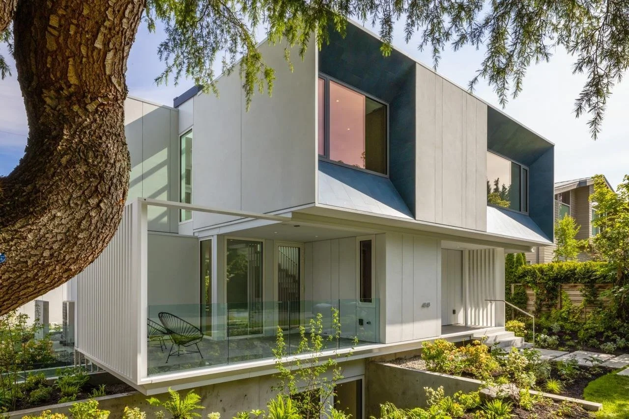 Modern two-story house with large windows, a balcony with glass railing, surrounded by greenery and trees.