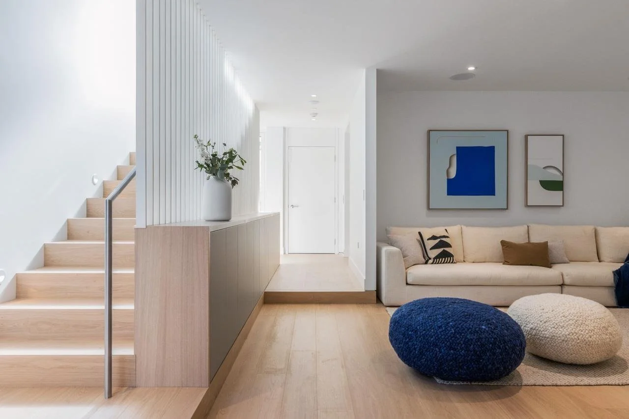 Modern living room with light wood flooring, white walls, a white sofa with beige, brown, and patterned pillows, two round poufs in navy blue and beige, and framed abstract artwork on the wall. A step-up hallway with a white railing and a vase with g