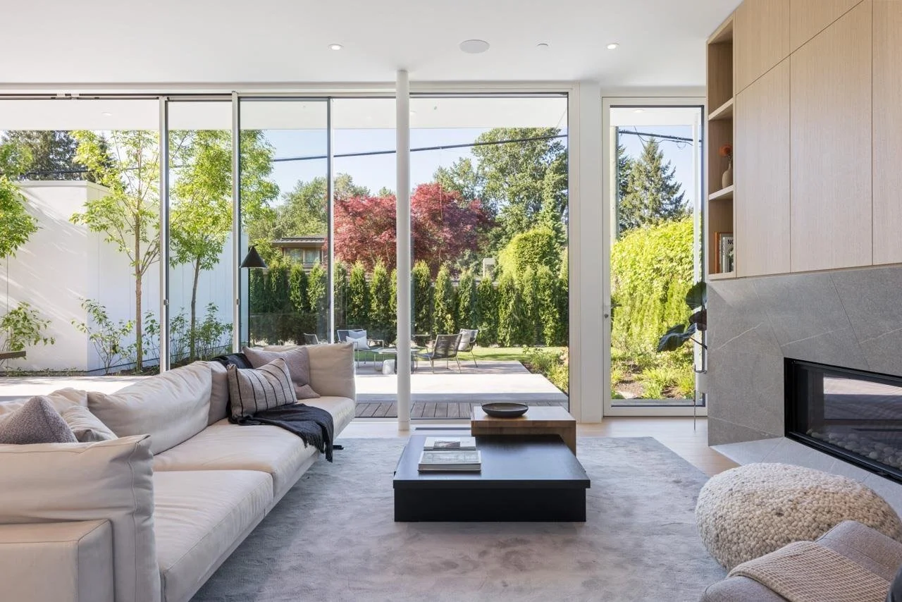 Living room with a large beige sectional sofa, black coffee table, and floor-to-ceiling glass doors leading to a green backyard with trees and outdoor seating.