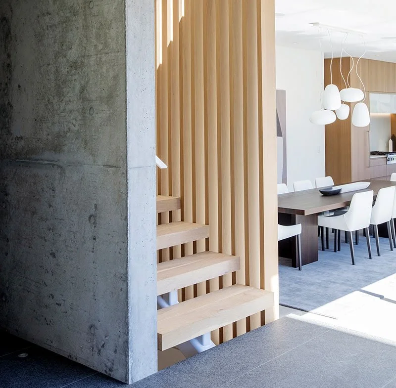 Interior of a modern home with a concrete wall, wooden slats, a staircase, and a dining area with a wooden table and white chairs.