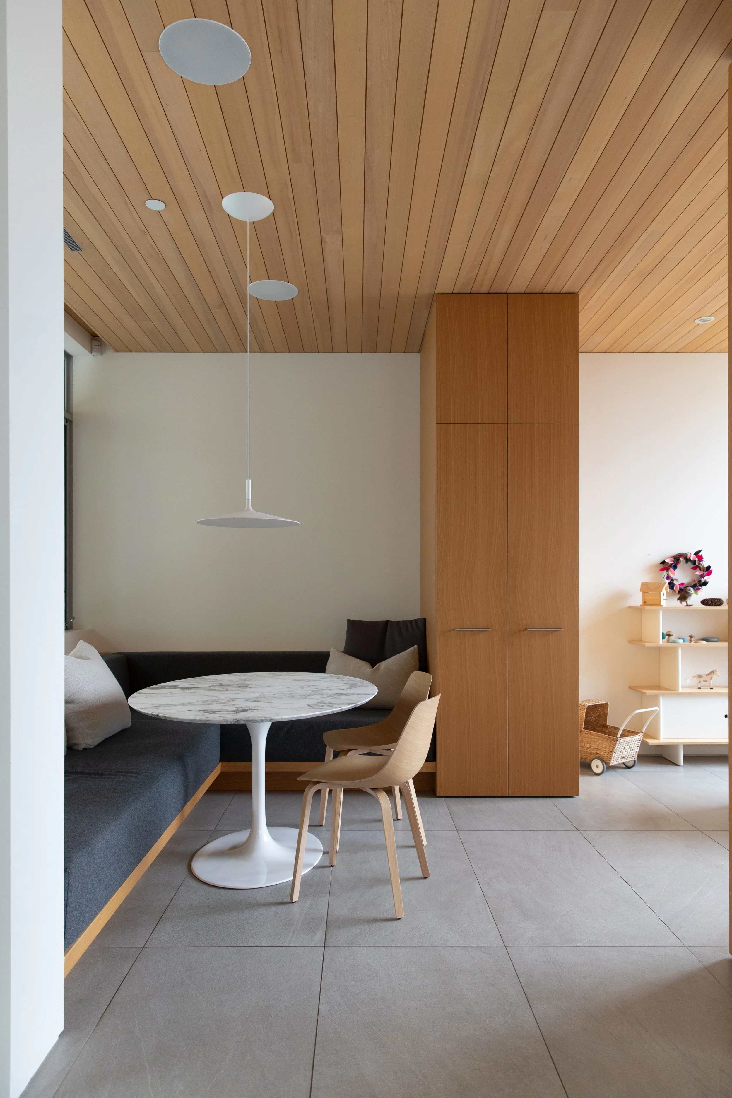 Interior of a minimalist dining area with a curved built-in bench, a round marble-topped table, two beige wooden chairs, a wooden cabinet, and a white shelf with small decorations, all under a wood-paneled ceiling.