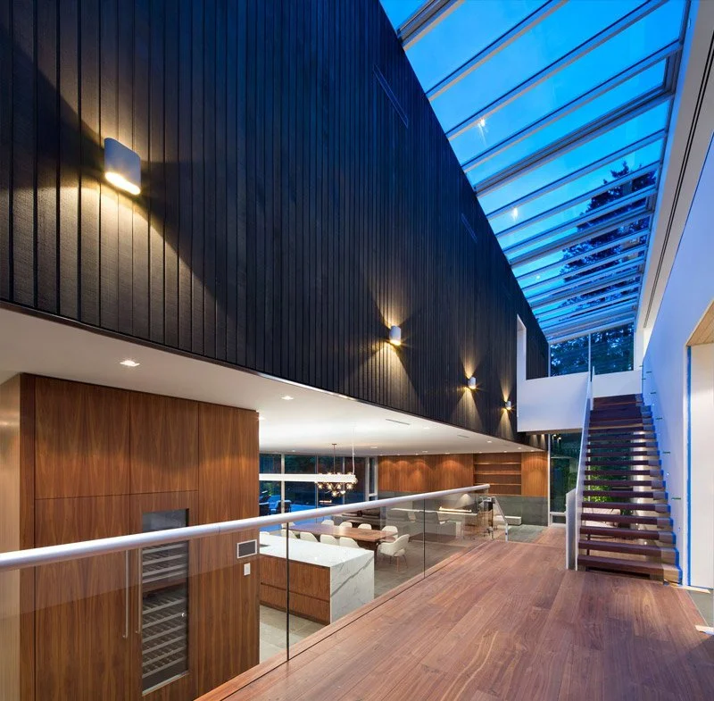 Modern interior with wooden flooring, glass railing, staircase, wood-paneled and black wall, and a large glass ceiling