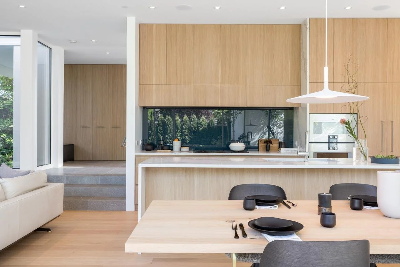 Modern open-concept kitchen and dining area with light wood cabinets, black dining ware, and a white countertop, adjacent to a living space with a beige sofa and large windows showing greenery outside.