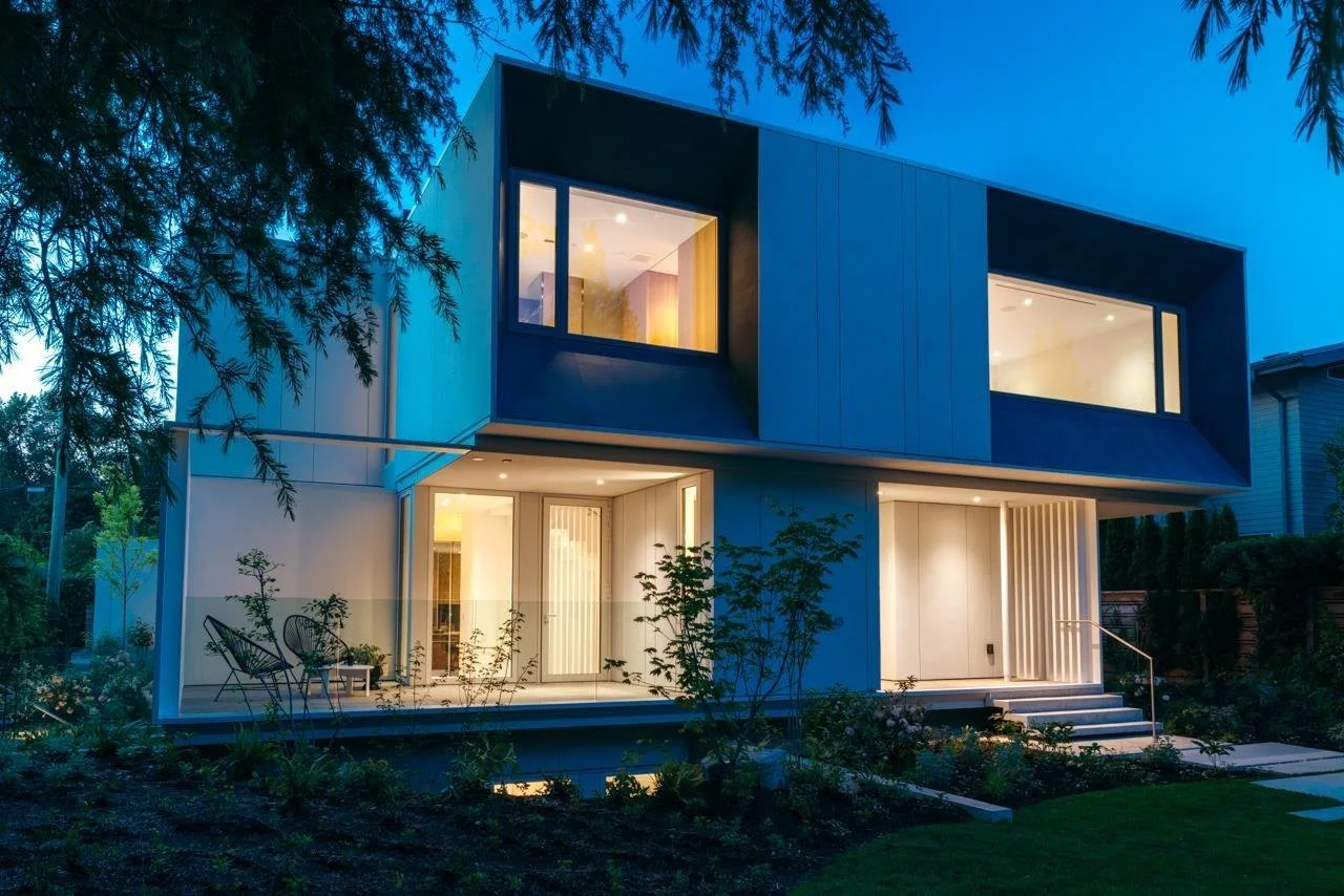 Modern two-story house with large windows, illuminated at dusk, surrounded by garden with trees and plants.