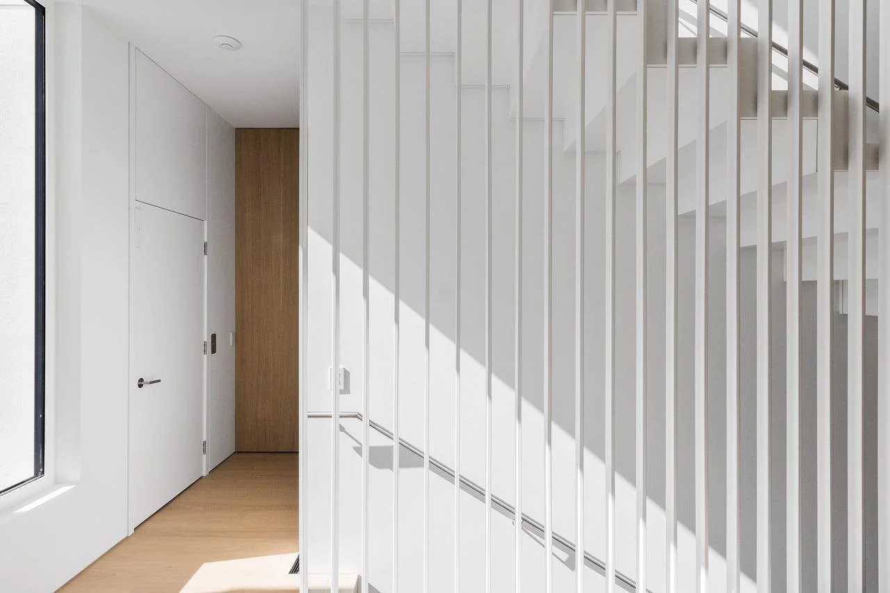 Interior view of a modern, minimal staircase with white vertical railings, white walls, wooden flooring, and a door at the bottom of the stairs.