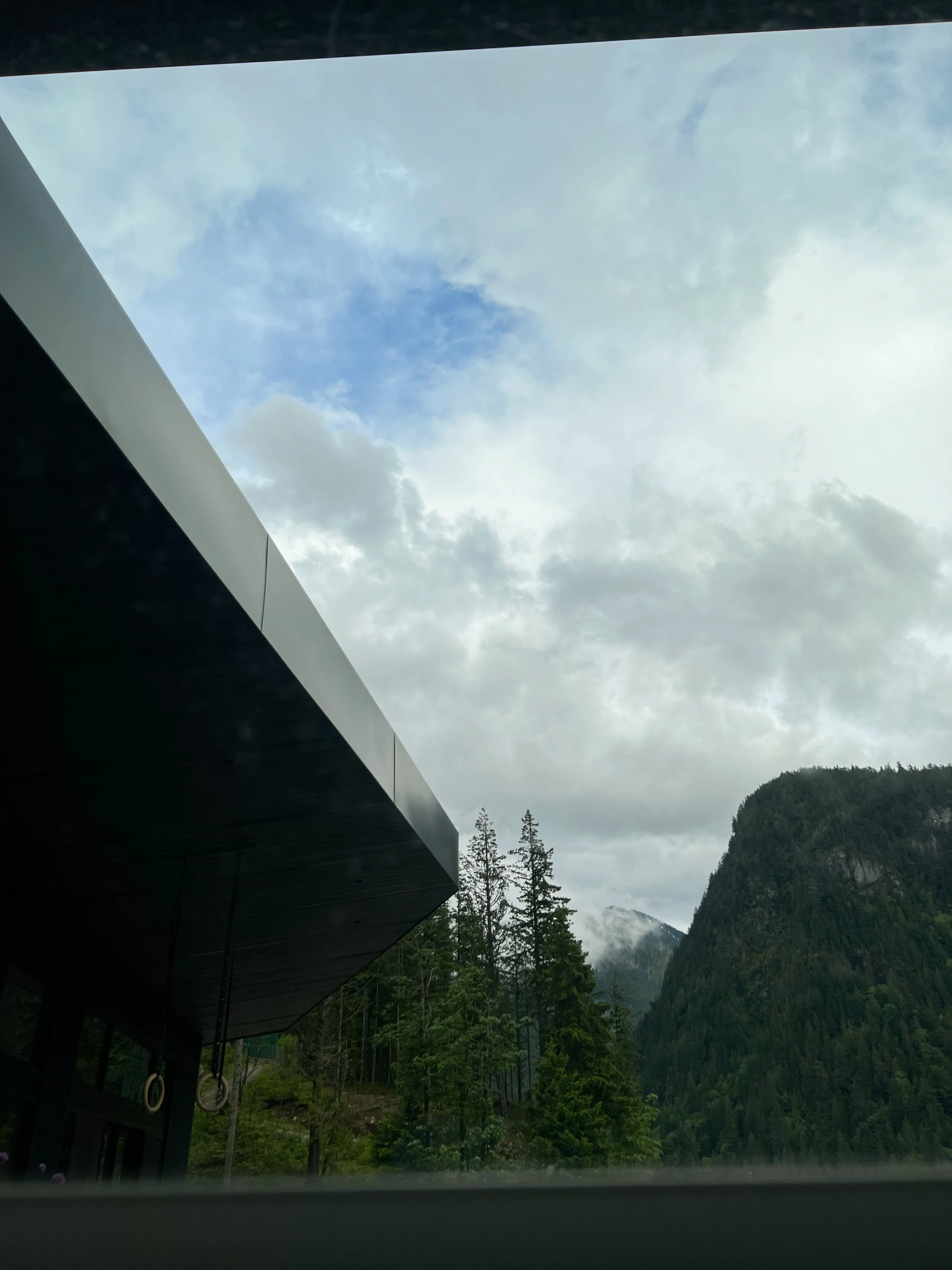 Cloudy sky with patches of blue, green forested mountain, and part of a modern building with rings hanging from the overhang.