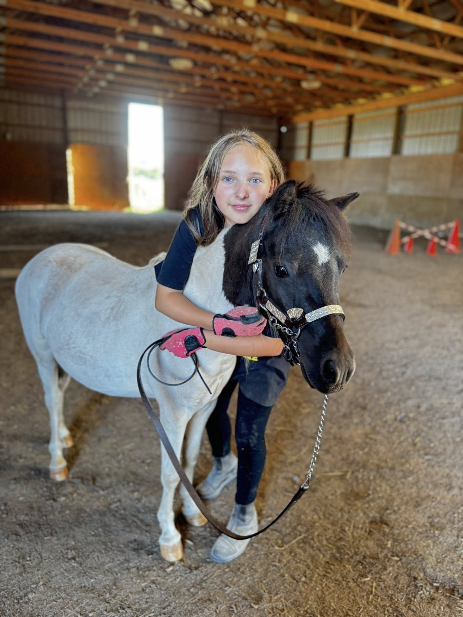 A young girl with blonde hair and blue eyes hugging a small black and white pony inside a barn.