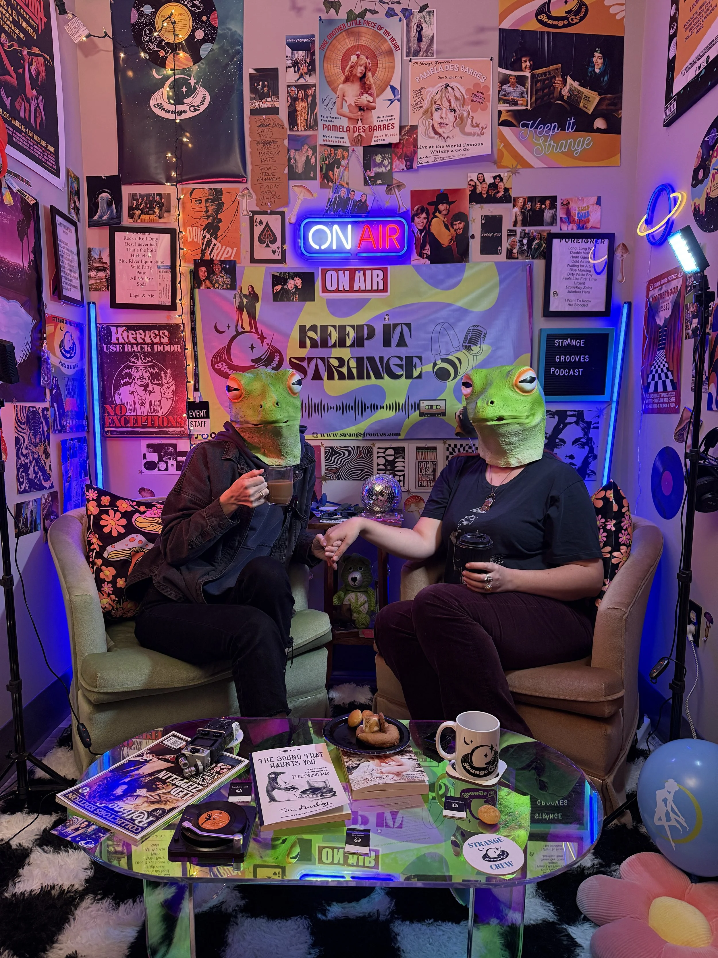 Two people with frog masks sitting on chairs in a room decorated with posters, neon signs, and colorfulLighting. The room has a table with books, magazines, and snacks, along with various retro and pop culture decor.