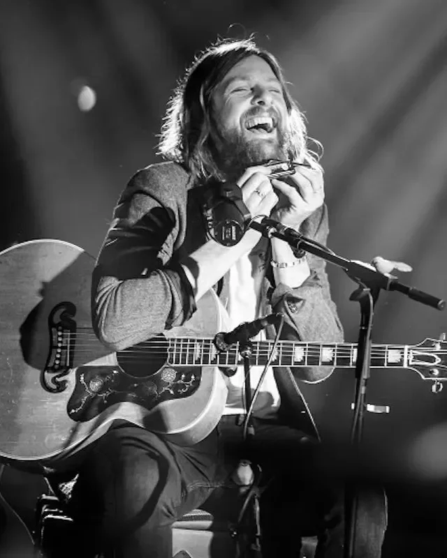 A man with long hair and a beard is smiling and laughing while sitting on a chair with an acoustic guitar on his lap, holding a harmonica near his mouth, with a microphone in front of him, on stage.