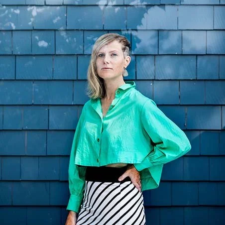 Woman standing in front of a blue-shingled wall, wearing a bright green long-sleeve shirt and black and white striped skirt.